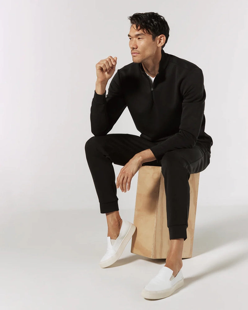 Man wearing black REV™ Jogger pants with white slip-on sneakers and a quarter-zip pullover, seated on wooden block – casual men's loungewear outfit