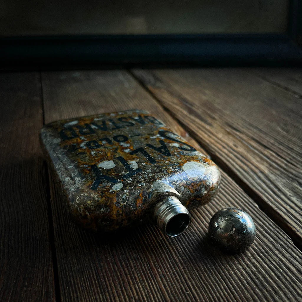 Salty Old Bastard etched stainless steel whiskey flask with patina finish resting on rustic wood table – The Simple Man barware