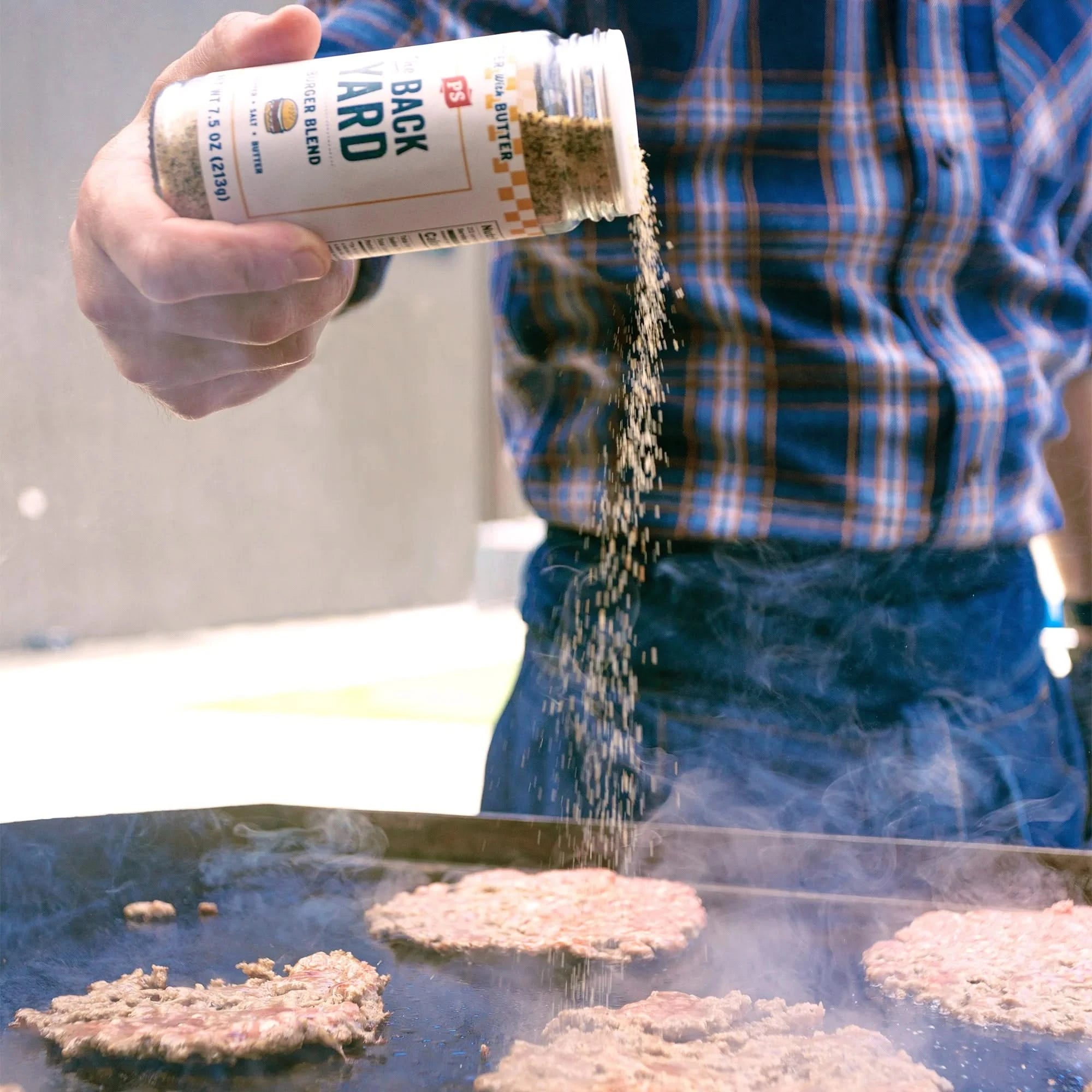 Man seasoning burgers on a flat-top grill using The Backyard Burger Blend from PS Seasoning, wearing a flannel shirt with steam rising in the background.