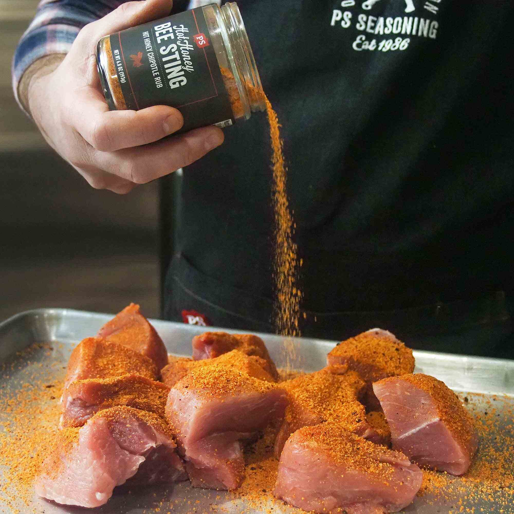 Sprinkling Hot Honey Bee Sting Rub on pork chunks – spicy chipotle BBQ rub with sweet heat being applied before grilling, featured at The Simple Man.
