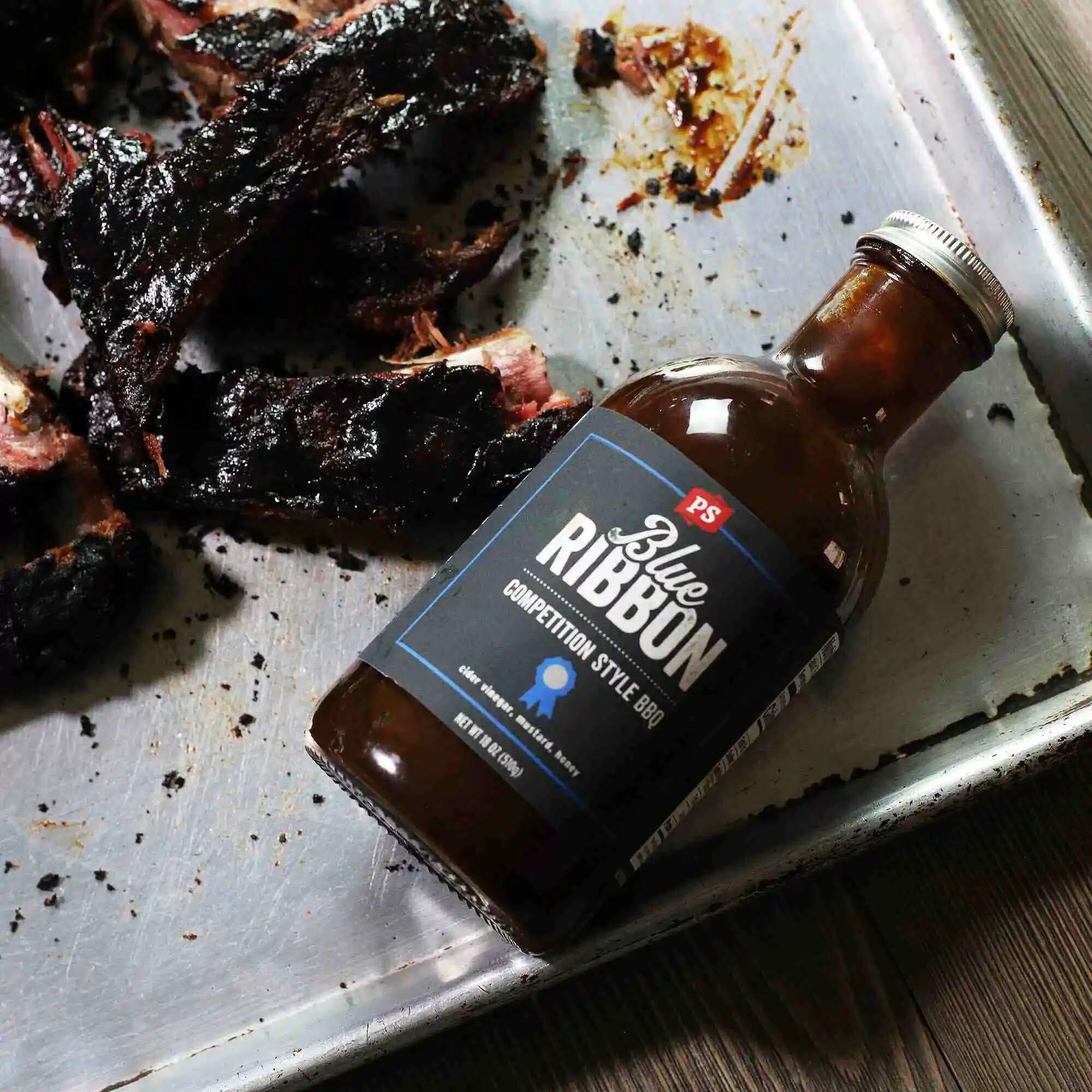 Blue Ribbon Competition Style BBQ Sauce bottle next to smoked ribs on a metal pan – perfect for backyard grilling and Kansas City-style barbecue flavor.