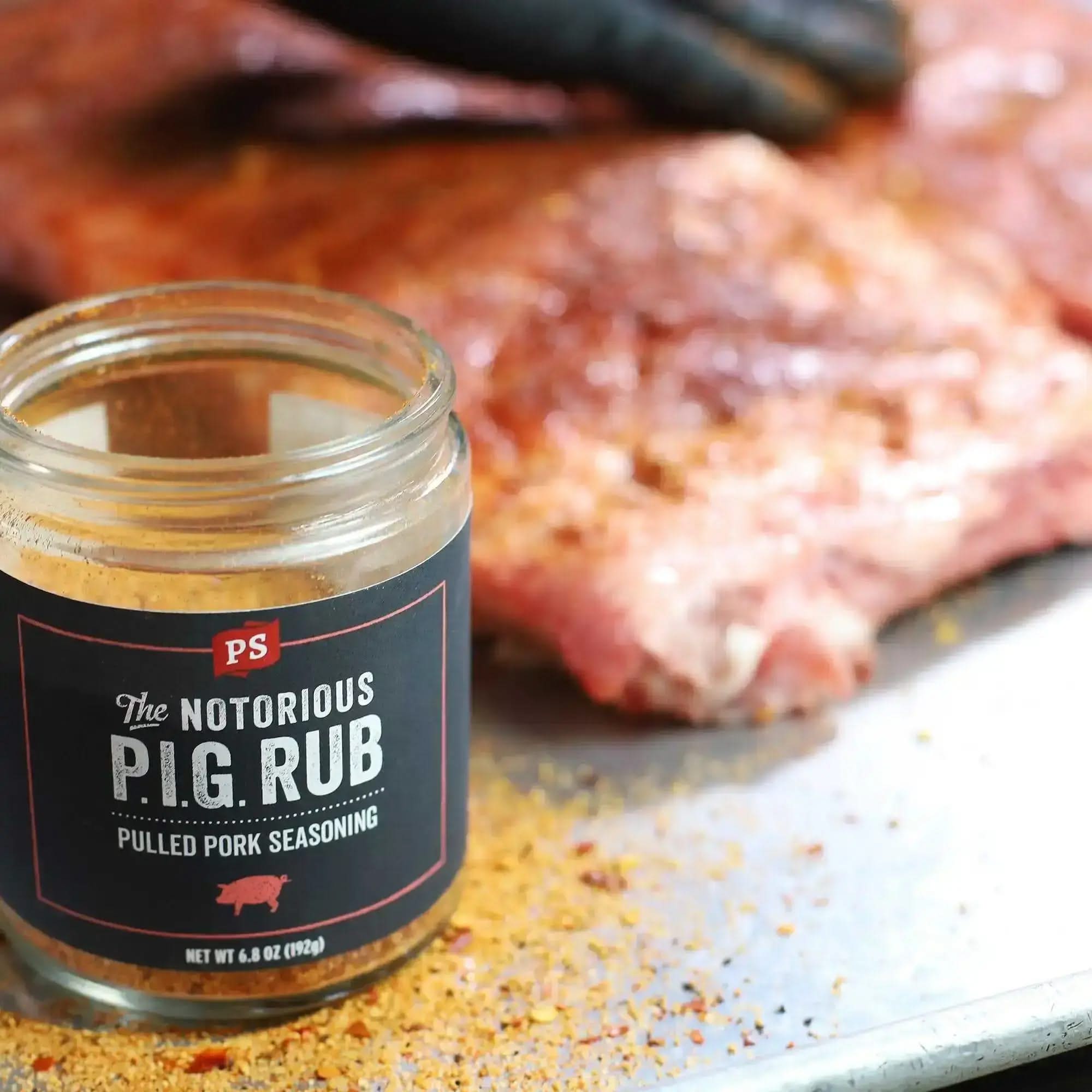 Jar of The Notorious P.I.G. Rub pulled pork seasoning beside seasoned pork ribs, showing texture and color of the spice blend on a metal tray.