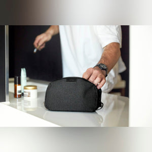 Bellroy Toiletry Kit in Black, on a bathroom counter with a hand on the zipper - The Simple Man