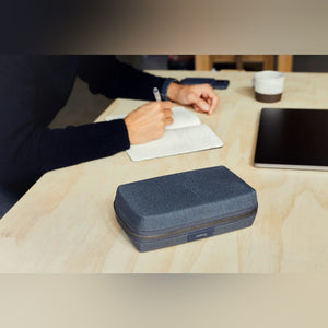 Bellroy Tech Kit cable organizer, shown in navy closed on a desk while a hand writes in a notebook - The Simple Man