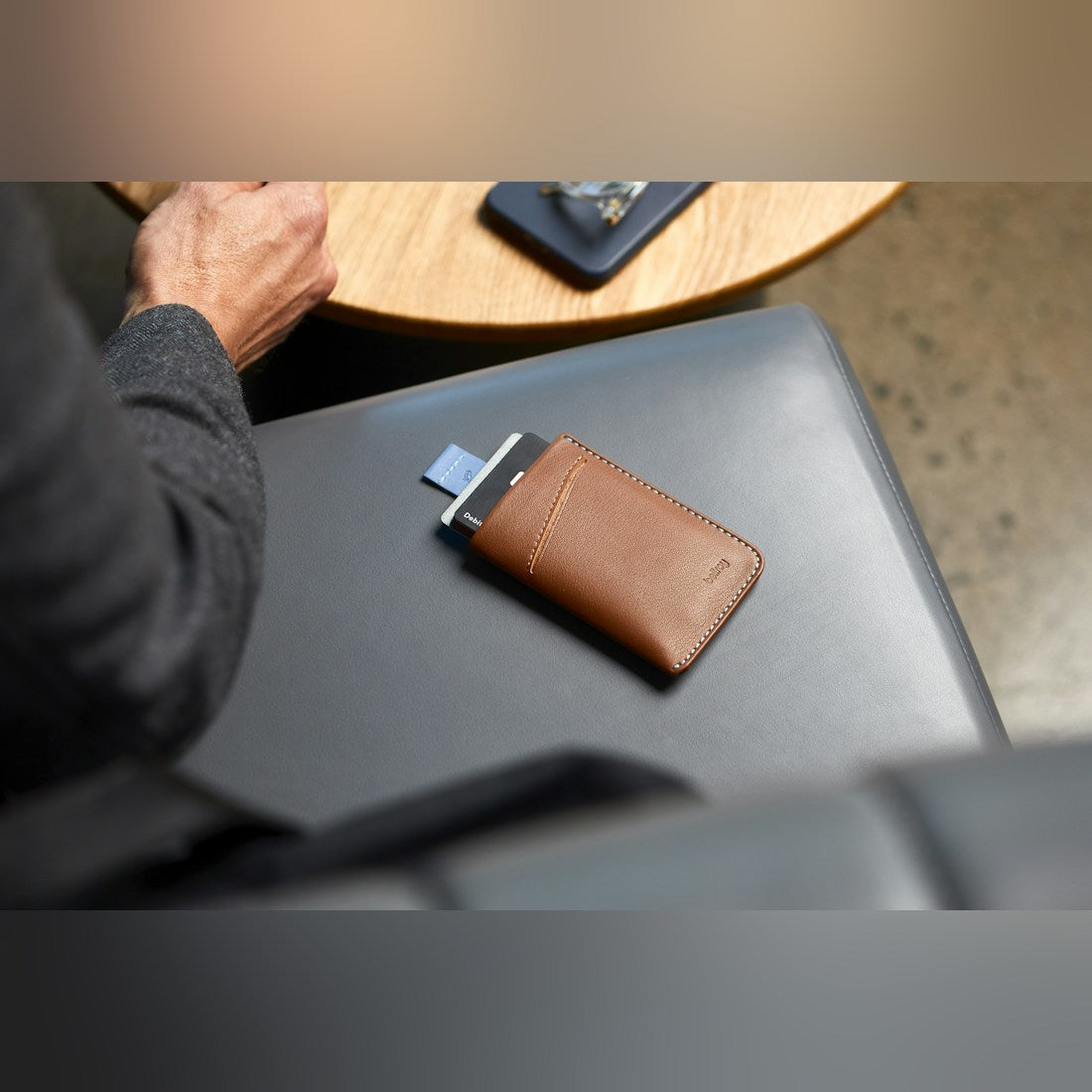 Bellroy Card Sleeve wallet, shown in tan leather with a blue pull tab, resting on a lounge seat - The Simple Man