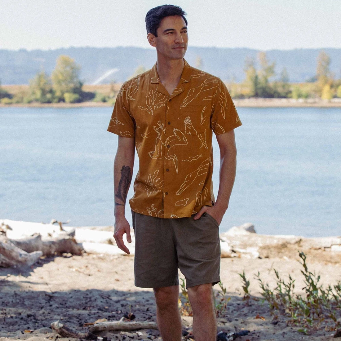 Man wearing Bridge and Burn Bodie Shirt Shoreflight amber on beach with driftwood — relaxed fit organic cotton camp collar men's summer shirt