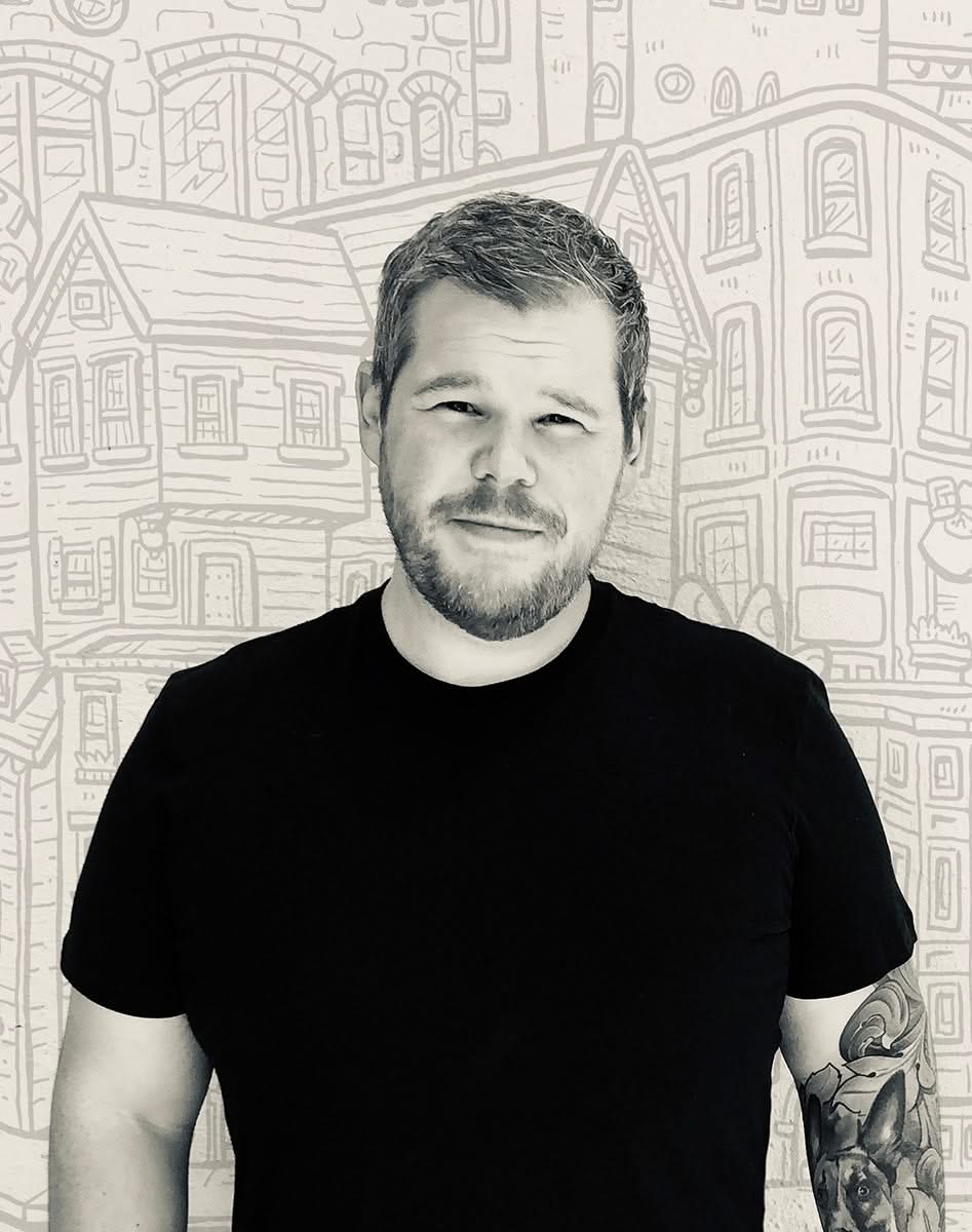 Artist Caleb Morris standing in front of his illustrated cityscape background, creator of the Omaha landmark print featured in The Simple Man's collection.