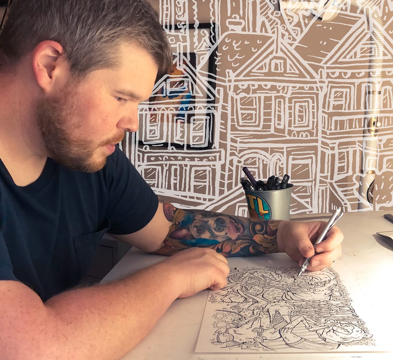 Illustrator Caleb Morris sketching a detailed city print at his desk, known for his “Welcome to the Neighborhood” series, including the Omaha artwork at The Simple Man.