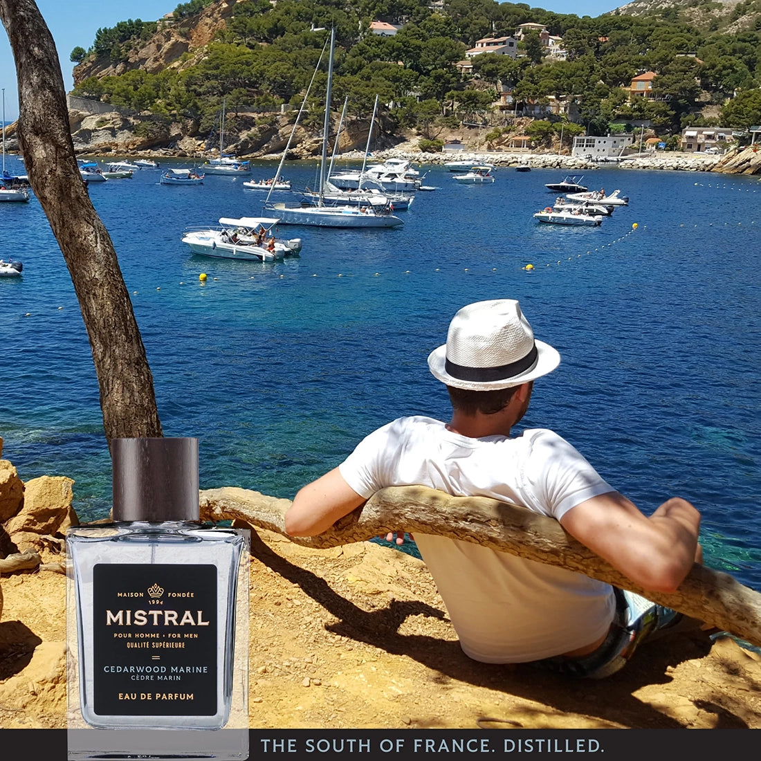 Mistral Cedarwood Marine Eau de Parfum bottle displayed in front of a coastal scene with a man overlooking blue water and sailboats.