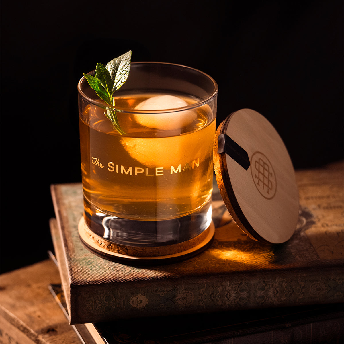 The Simple Man candle vessel becomes a  cocktail glass with whiskey, ice sphere, and sage garnish on vintage books with wood cork lid that becomes the coaster– The Simple Man
