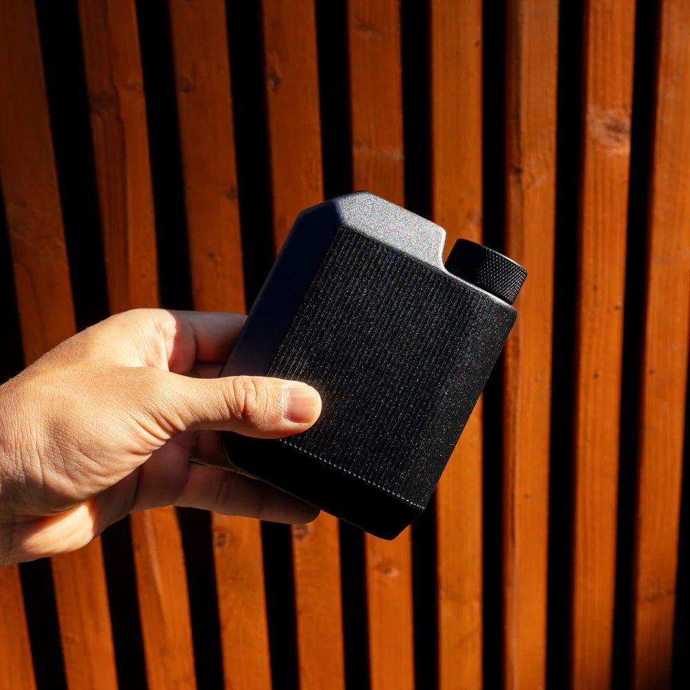 Man holding matte black Cool Flask outdoors against wooden background – slim stainless steel pocket flask from The Simple Man.