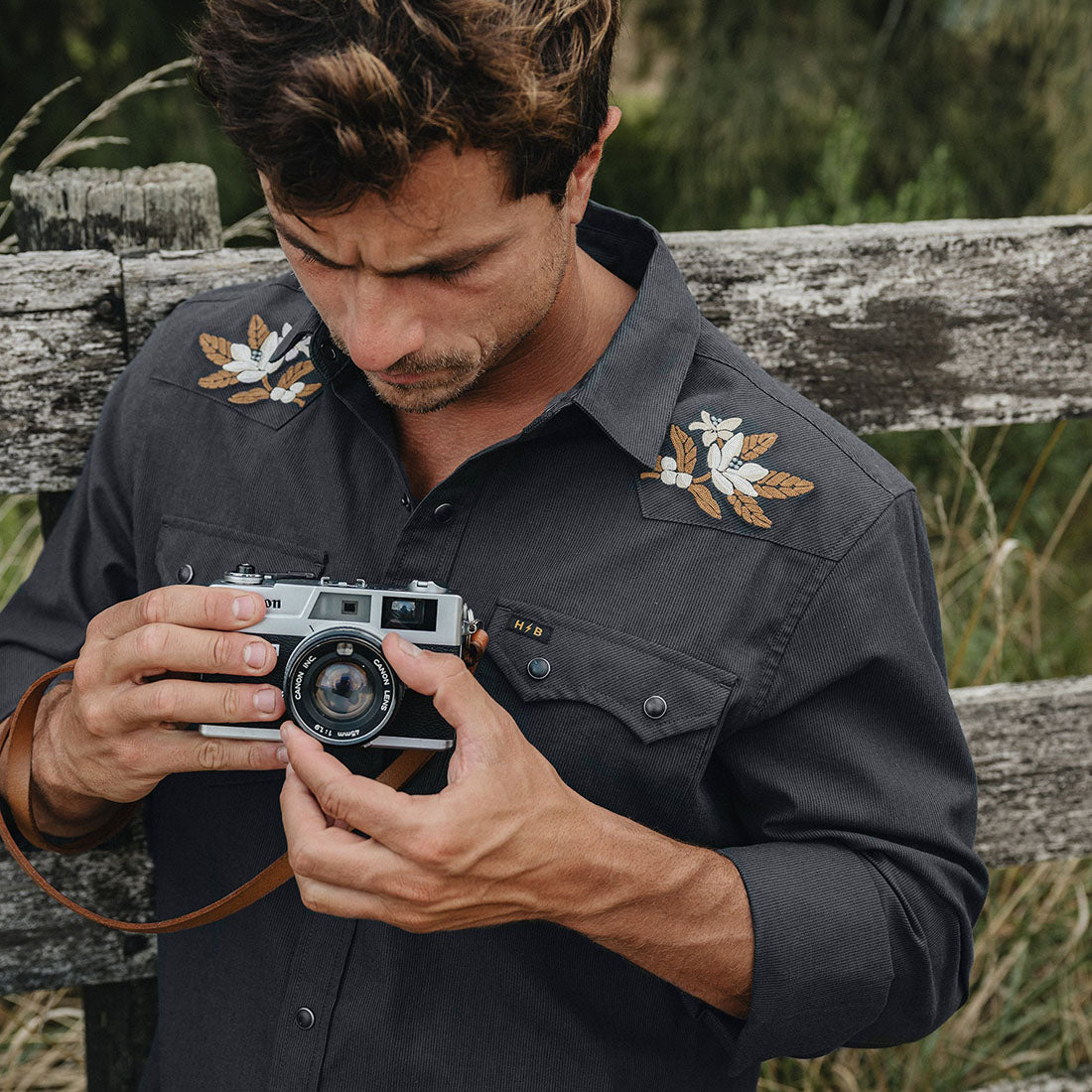 Man holding a camera while wearing the black Orange Blossoms Crosscut Deluxe Longsleeve, embroidered floral shoulder detail lifestyle shot -- The Simple Man