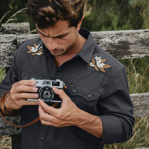 Man holding a camera while wearing the black Orange Blossoms Crosscut Deluxe Longsleeve, embroidered floral shoulder detail lifestyle shot -- The Simple Man