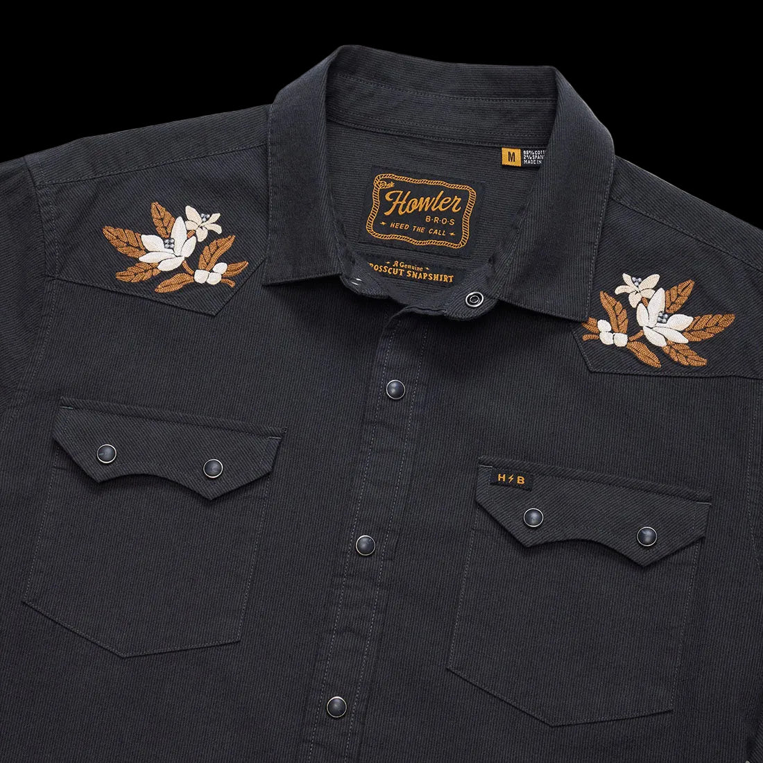 Close-up of embroidered floral shoulder yoke and pearl snap pockets on the black Orange Blossoms Crosscut Deluxe Longsleeve showing the Howler Bros label -- The Simple Man
