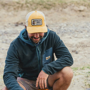 Man wearing the yellow corduroy Howler Brothers El Monito Surfs Badge Hat while sitting outdoors in casual fleece jacket.