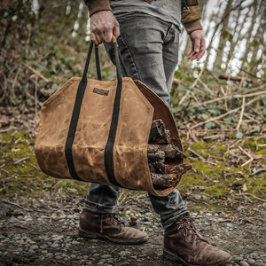 Man carrying Readywares waxed canvas firewood log carrier loaded with wood on gravel path outdoors — brown work boots lifestyle shot