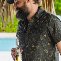 Man wearing Howler Bros H Bar B Snapshirt Sea Rodeo in outdoor tropical setting – gold charcoal western print pearl snap short sleeve shirt