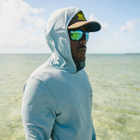 Man wearing Howler Bros HB Tech Hoodie pool blue with hood up by ocean – UPF 50+ lightweight sun protection fishing hoodie outdoor lifestyle

