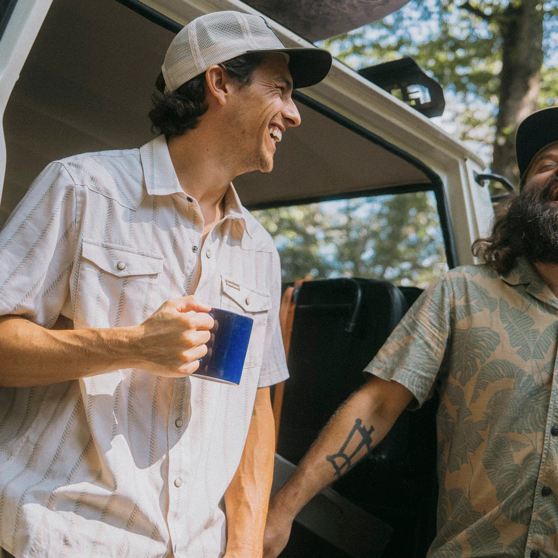 Man wearing Howler Brothers H Bar B Snapshirt Land Cruiser edition laughing outdoors near camper van with coffee mug