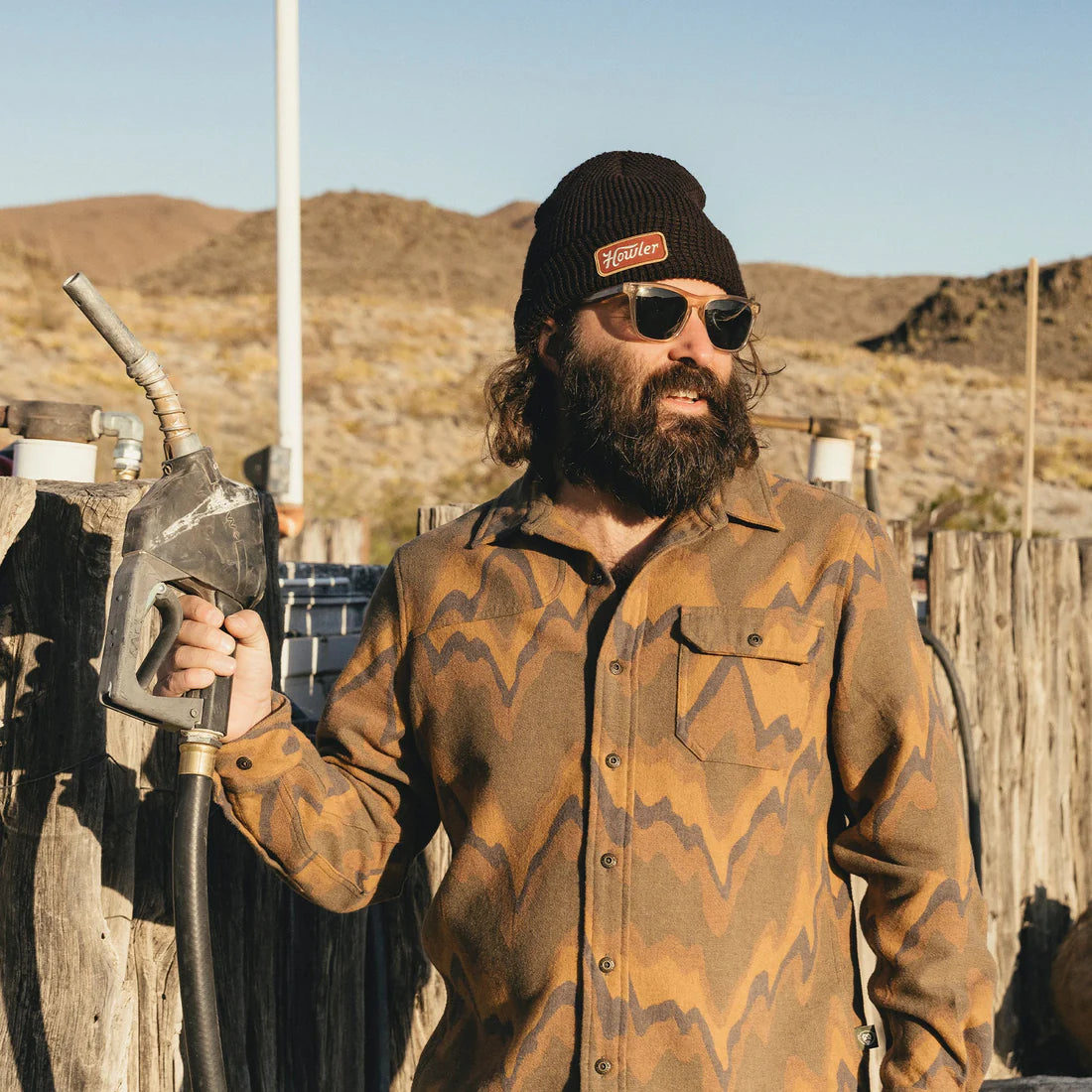 Man wearing Hawkeye Beanie by Howler Brothers, holding gas pump in rugged desert setting with patterned shirt