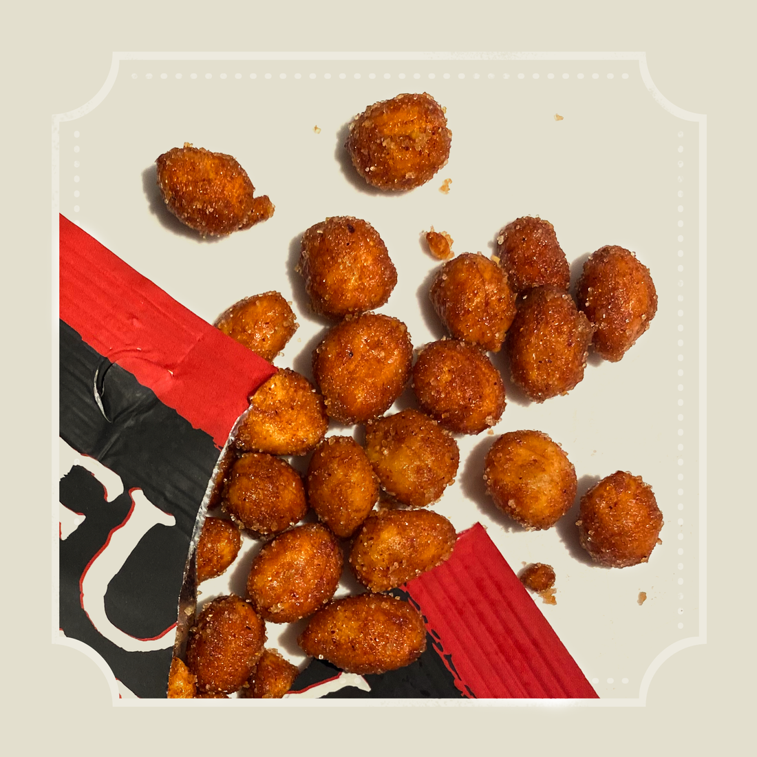 Glazed hot BBQ peanuts spilling from a torn red and black packet onto a cream backdrop