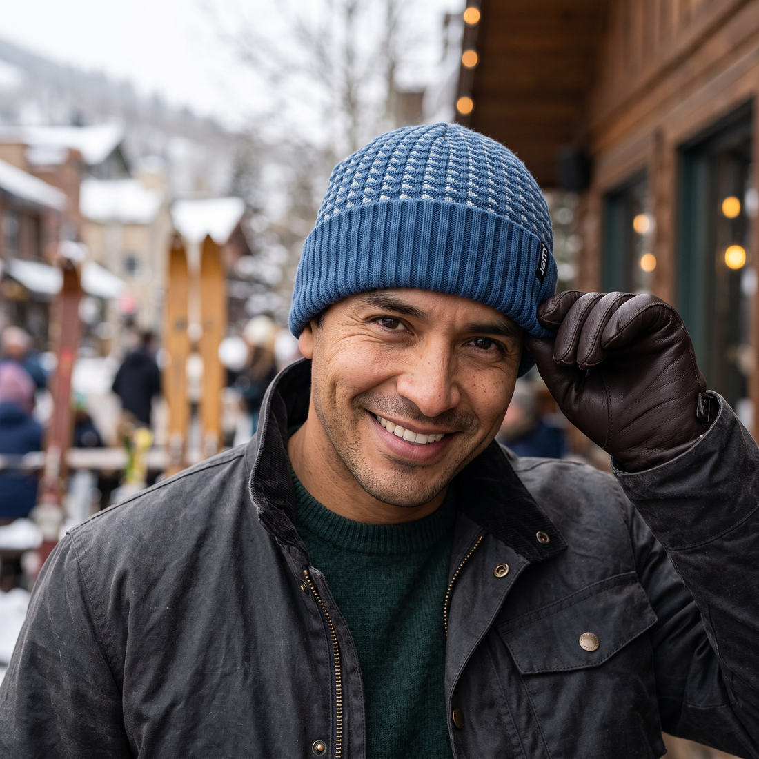 Smiling man in a blue Huey Beanie with a pale pattern crown, tipping the cuff with a leather glove on a snowy street
