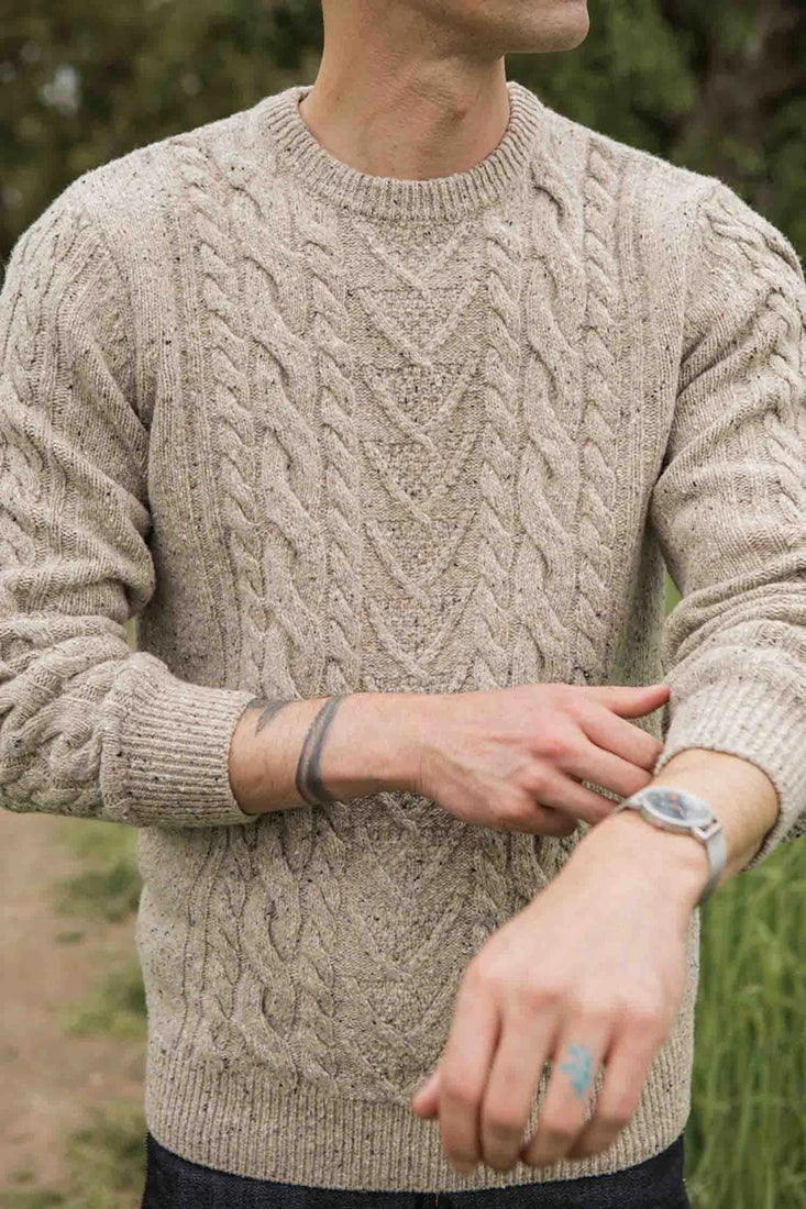 Man wearing The Ludlow Sweater by Bridge & Burn in oatmeal cable knit, styled casually with jeans outdoors