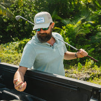 Man wearing Howler Brothers Pilgrimage Polo in Riviera Blue with Howler Bros trucker hat – men's lifestyle polo shirt