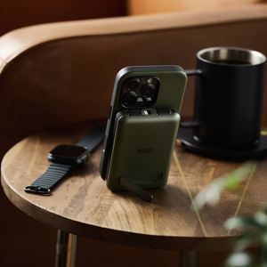 Matte Olive power bank propped on its kickstand behind a phone, mug and watch on a wood table