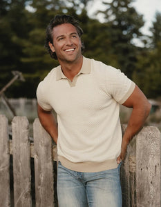 Man wearing The Normal Brand Robles Polo outdoors leaning on fence – ivory khaki knit jacquard men's polo casual lifestyle short sleeve