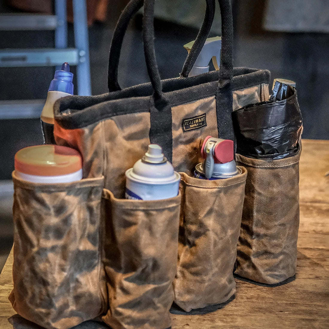 Readywares Supply Bag loaded with spray cans cleaning supplies and tools on workbench — 14-pocket waxed canvas utility tote in workshop