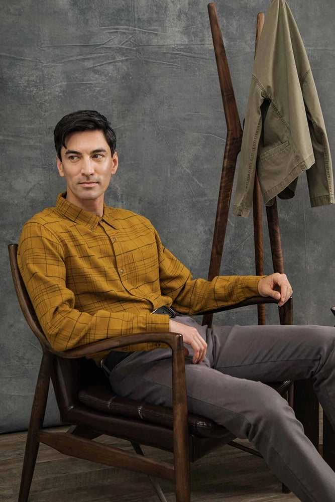 Model wearing Sutton Slim Shirt in Golden Horizon mustard plaid with grey chinos, seated in a chair