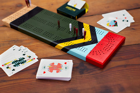 Painted cherry wood cribbage board in play with pegs and playing cards on a rustic tabletop.