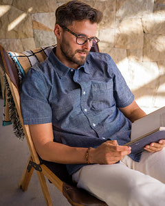 Man wearing Ace Rivington Indigo Rinse chambray short sleeve shirt seated outdoors reading — slim fit men's chambray button-down