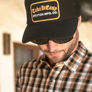 Man wearing Brixton Take It Easy Washed Black trucker hat with plaid shirt and sunglasses — gold patch old English script men's snapback