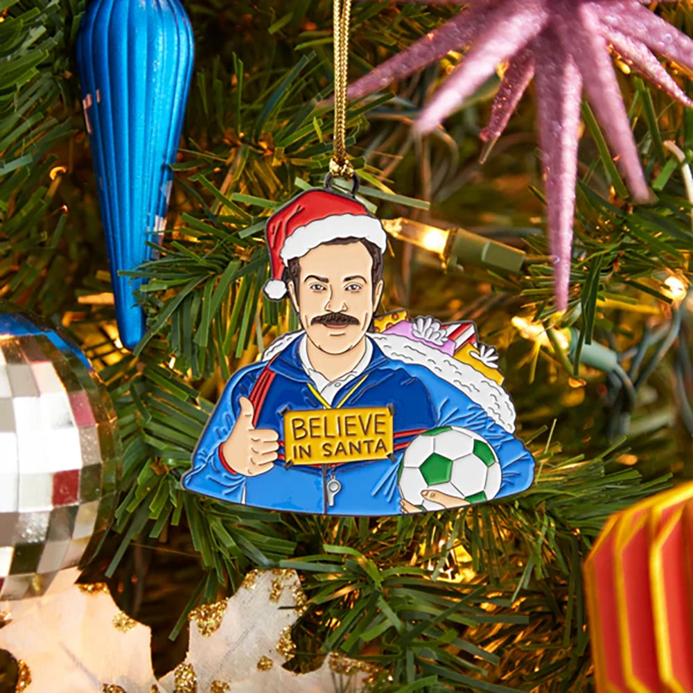 Hand-illustrated Ted “Believe in Santa” enamel ornament hanging on a Christmas tree with holiday décor.