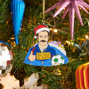Hand-illustrated Ted “Believe in Santa” enamel ornament hanging on a Christmas tree with holiday décor.