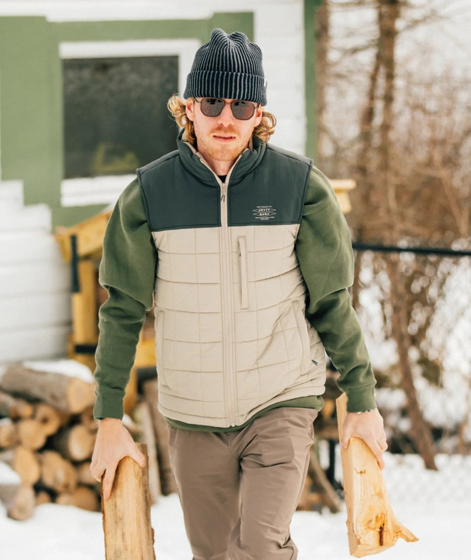 Man wearing Jetty Terrace Vest in Oyster color while carrying firewood outdoors in winter – available at The Simple Man men’s clothing store in Omaha.