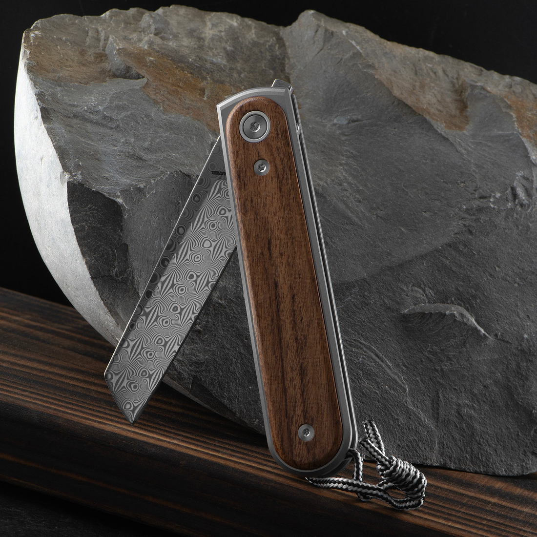 Duval rosewood pocket knife with Damasteel blade half open, leaning on slate with lanyard
