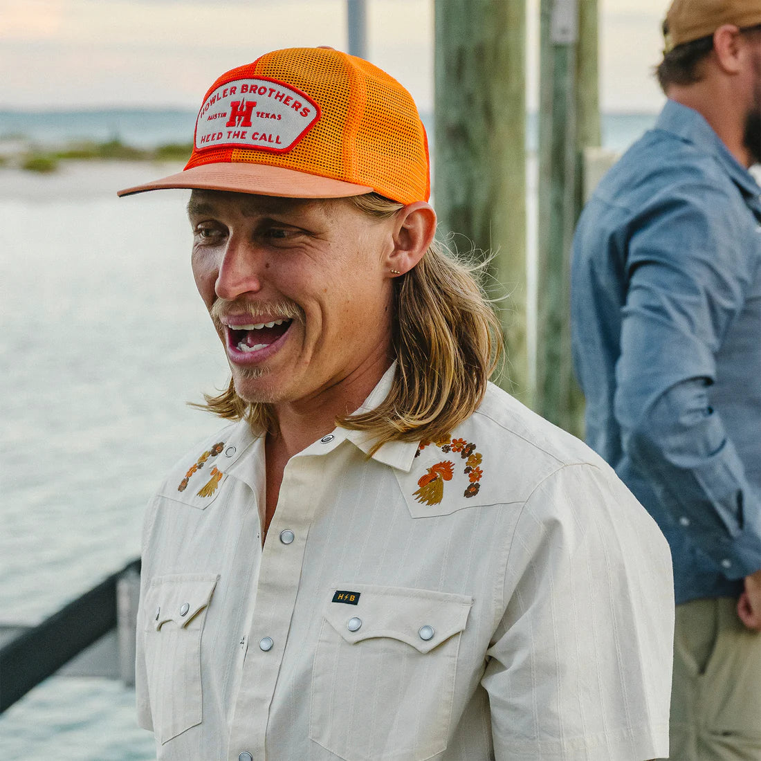 Man wearing Howler Brothers Feedstore Snapback in orange at waterfront dock – men's mesh trucker hat with Heed the Call patch