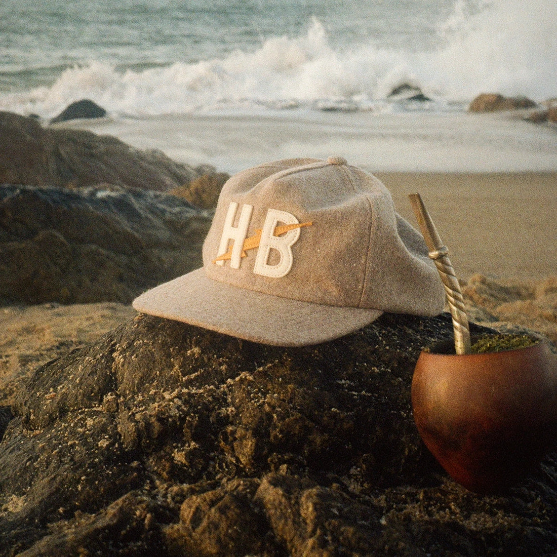 Big HB Unstructured Snapback Hat in Cowhide resting on a beach rock, tan wool cap with HB lightning bolt patch, lifestyle shot -- The Simple Man