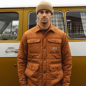 Man in the toffee Windward puffer jacket and a tan beanie standing in front of a vintage yellow van
