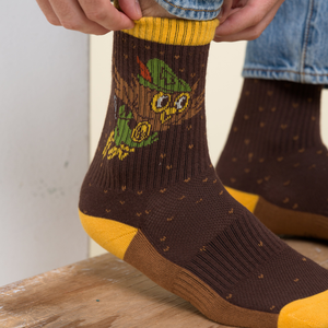 Lifestyle shot of a man pulling up a brown Woodsy Owl crew sock over a mustard heel