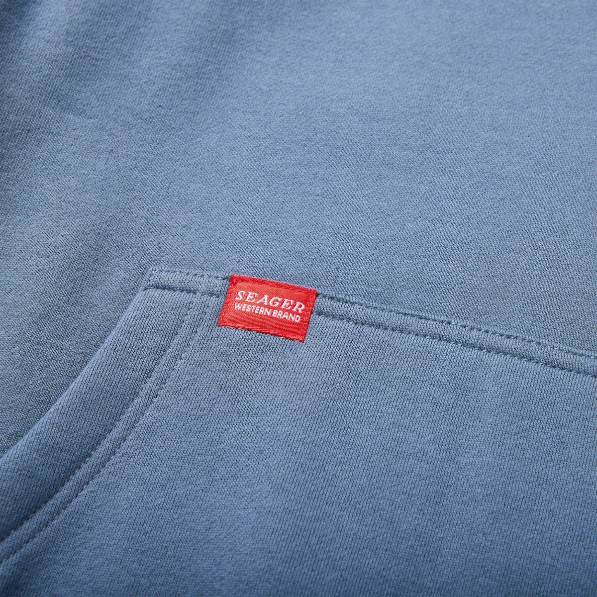 Seager Company hoodie in petrol blue featuring red Western Brand tag above front kangaroo pocket – rugged men’s hoodie detail.