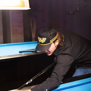 Man wearing Seager x Waylon Jennings Flying W Snapback in black and yellow while playing pool in a dimly lit bar setting