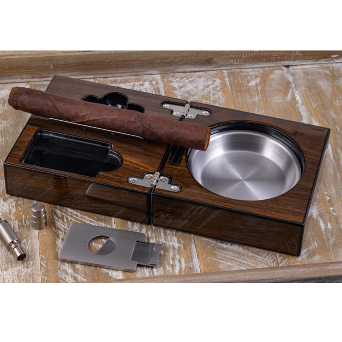 Folding walnut cigar ashtray set open with cigar resting — stainless steel ash reservoir guillotine cutter and punch cutter on wood surface