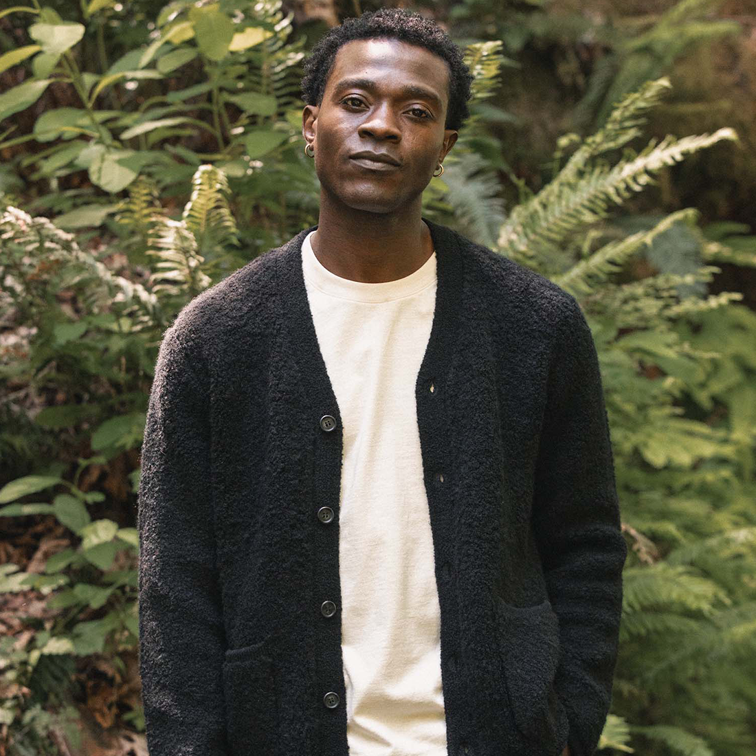 Man wearing the Mateo cardigan in black over a cream tee among ferns, front view - The Simple Man