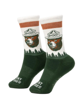 Prevent Wildfires Sock in Spruce lifestyle shot feet up on ottoman by fireplace in cabin with hiking boots showing Smokey Bear knit design