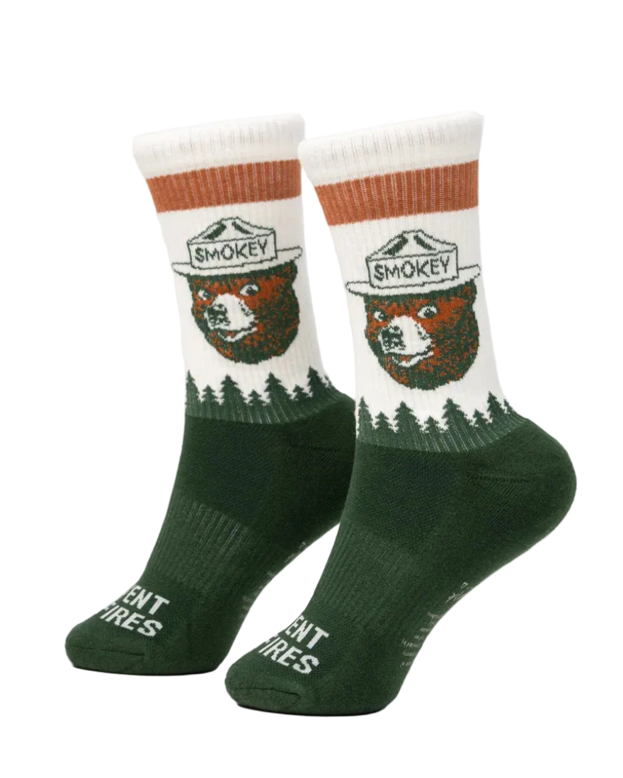 Prevent Wildfires Sock in Spruce lifestyle shot feet up on ottoman by fireplace in cabin with hiking boots showing Smokey Bear knit design