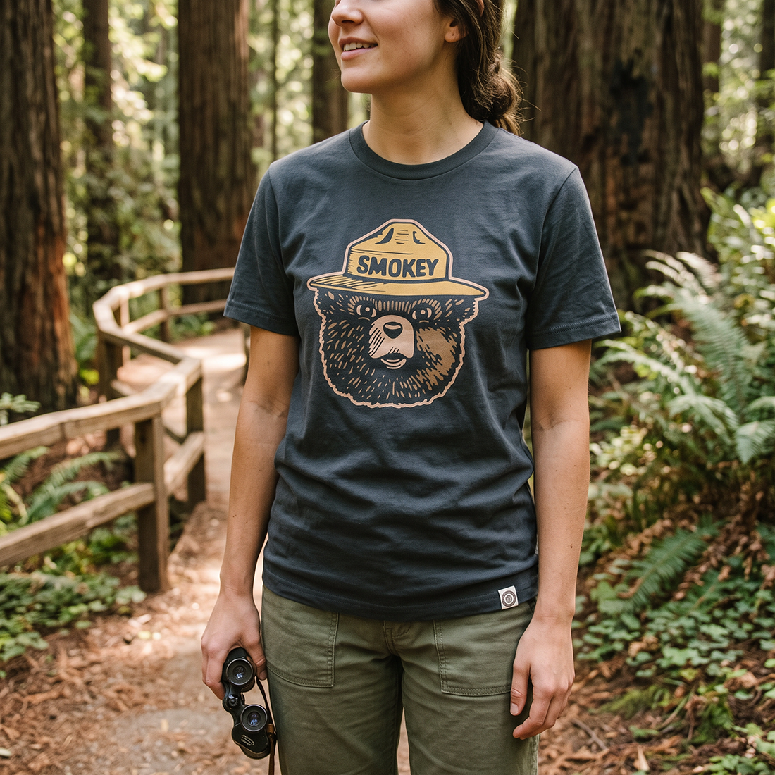 Smokey Bear Logo Tee in Dark Navy worn by woman on forest trail holding binoculars by The Landmark Project